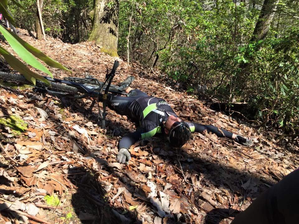 A mountain biker lies on the ground beside their bike, surrounded by a forested area covered in leaves. The cyclist is wearing a black and green outfit and appears to be resting or recovering after falling. Big South Fork mountain bike trail.