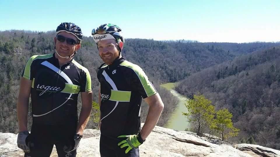 Two male cyclists standing on a rocky overlook, wearing matching black and green cycling jerseys and helmets. Behind them, a scenic view of a river winding through a forested valley under a clear blue sky. The sun is shining, highlighting the natural beauty of the landscape. Big South Fork mountain bike trail.