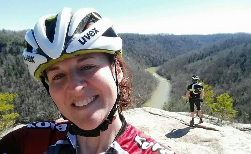 A woman in a cycling helmet smiles for a selfie while standing on a rocky overlook. In the background, another cyclist gazes out at a winding river and a landscape of trees under a clear blue sky. The scene captures a moment of outdoor adventure and camaraderie in nature. Big South Fork mountain bike trail.
