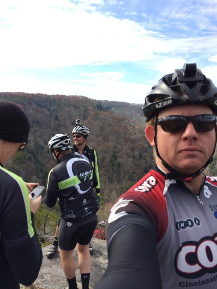 A group of three cyclists paused at a scenic overlook, surrounded by a colorful autumn landscape. One cyclist is taking a selfie, while the others engage in conversation, with one checking a phone. They are dressed in cycling gear and helmets, and the sky is clear with scattered clouds. Big South Fork mountain bike trail.