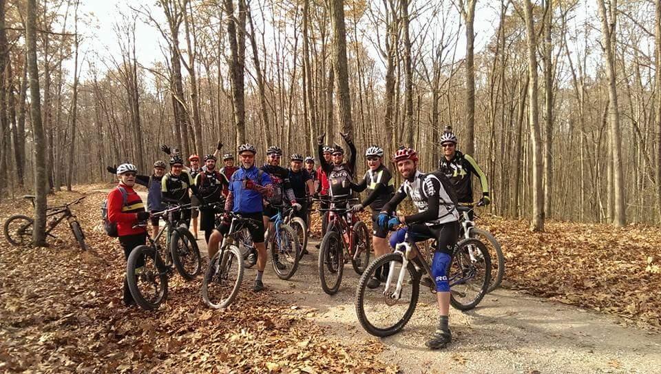 Group of mountain bikers gathered on a wooded trail, surrounded by trees with autumn leaves. They are all wearing helmets and various cycling attire, some holding their bikes while others smile at the camera. Big South Fork mountain bike trail.