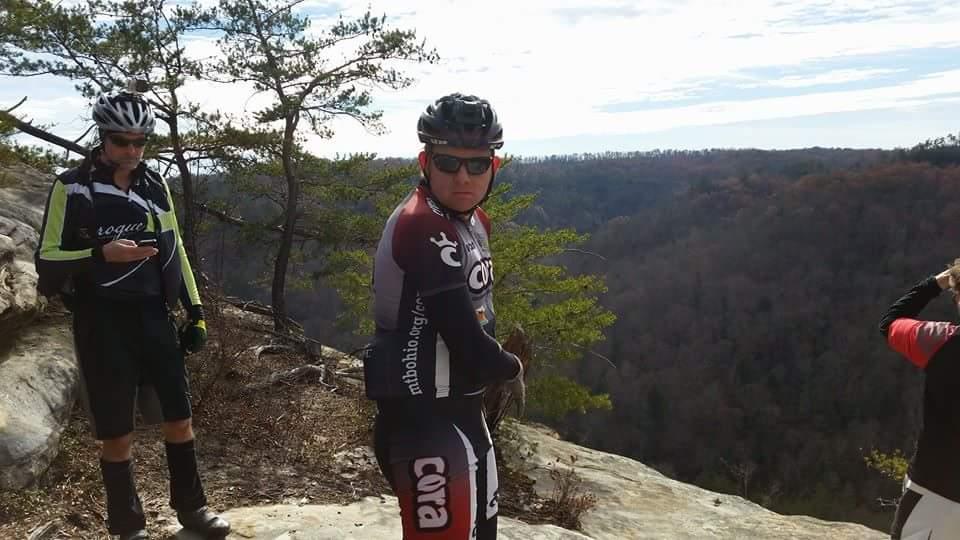 Two mountain bikers stand on a rocky ledge overlooking a scenic valley. One cyclist, wearing a black and red jersey with sunglasses and a helmet, faces the camera while the other, in a black and green jersey, checks their phone. Trees frame the background, and a cloudy sky stretches above. The landscape showcases rolling hills with autumn foliage. Big South Fork mountain bike trail.
