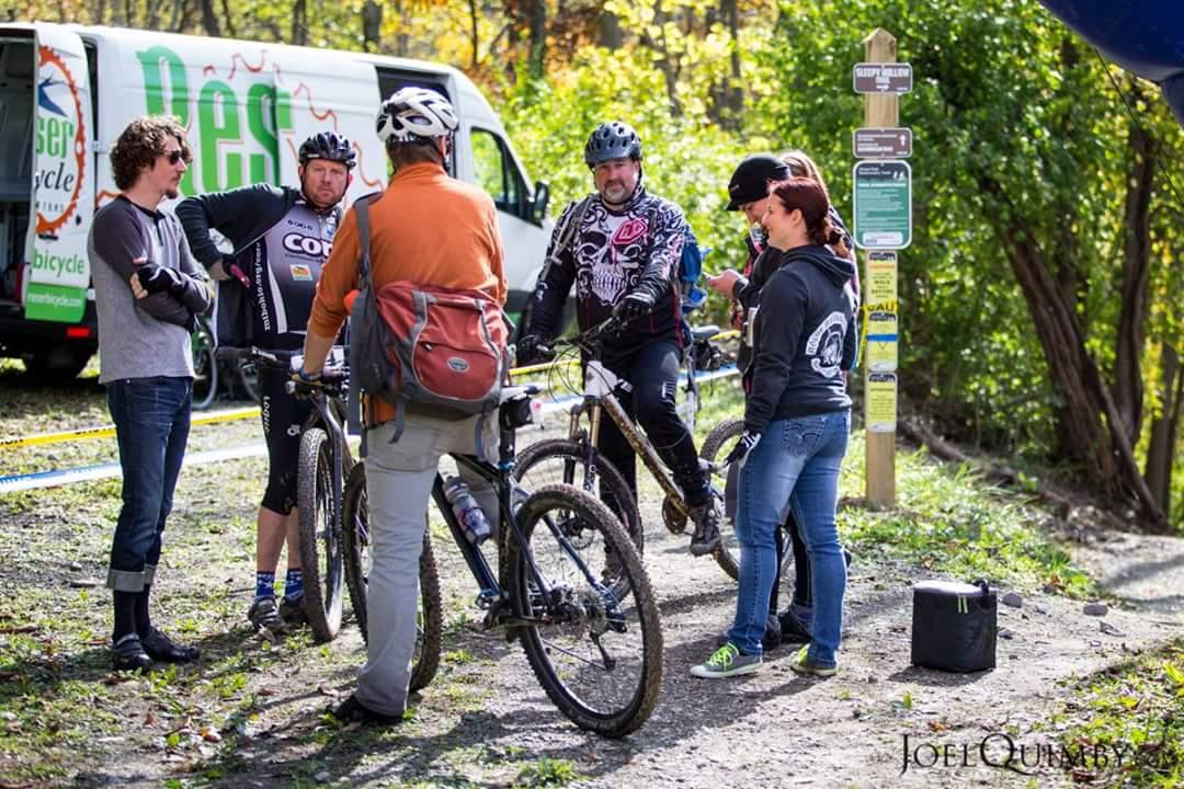 A group of cyclists gathered near their mountain bikes, engaging in conversation at a trailhead. The background features a vehicle with a bicycle logo and trail signage, surrounded by autumn foliage. Devou Park mountain bike trail.