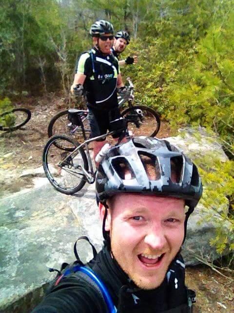 A group of three mountain bikers posing for a selfie on a rocky outcrop in a lush, green outdoor setting. Two bikers are in the background, smiling and making playful gestures, while the front biker is smiling widely at the camera. All are wearing helmets and cycling gear. Big South Fork mountain bike trail.