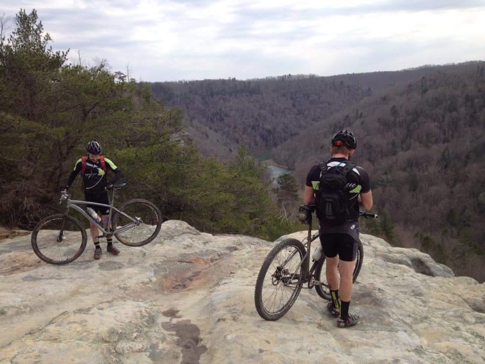 Two mountain bikers pause on a rocky overlook, each with their bikes. In the background, a scenic valley with trees and distant hills extends towards a winding river, under a cloudy sky. The terrain appears rugged, showcasing a mix of natural beauty and outdoor adventure. Big South Fork mountain bike trail.