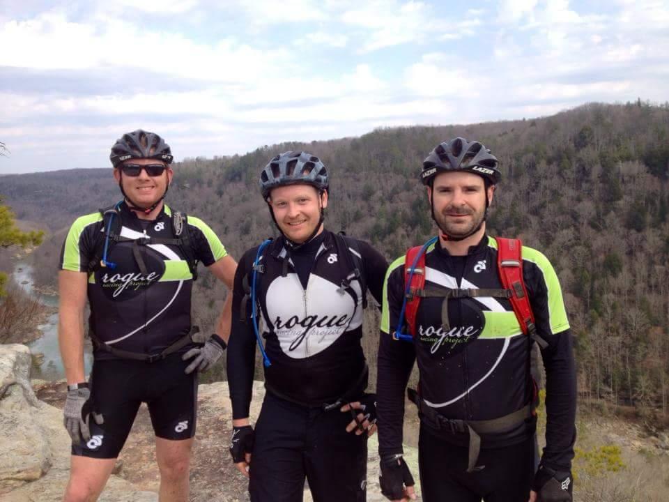 Three male cyclists wearing matching black and green jerseys and helmets stand on a rocky outcrop with a forested valley in the background. They are smiling and engaged in outdoor recreational activities. The sky is slightly overcast, indicating a cool day for biking. Big South Fork mountain bike trail.