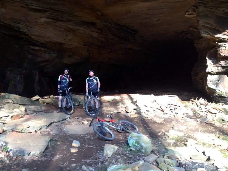 Two mountain bikers standing next to their bikes in a rocky area near a cave entrance. The background features a large rock formation, and sunlight is filtering into the cave, highlighting the ground covered with stones and foliage. Big South Fork mountain bike trail.