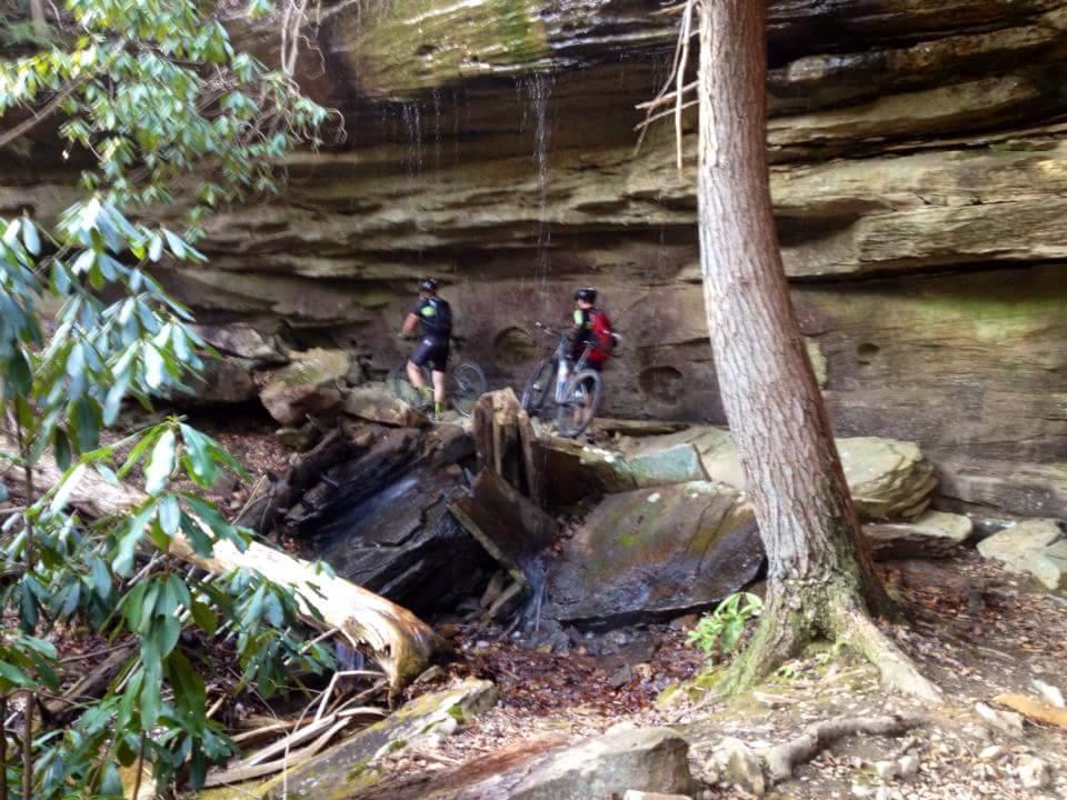 Two mountain bikers stand beside their bikes in a forested area with rocky cliffs and dripping water. Dense greenery surrounds the scene, and fallen logs are scattered on the ground, creating a natural and adventurous atmosphere. Big South Fork mountain bike trail.
