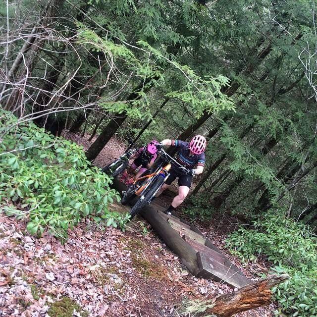 A mountain biker navigating over a log on a forest trail, surrounded by greenery and trees. The rider is wearing a helmet with pink accents and is focused on overcoming the obstacle. A second bike is visible nearby, suggesting a companion or support on the ride. Big South Fork mountain bike trail.