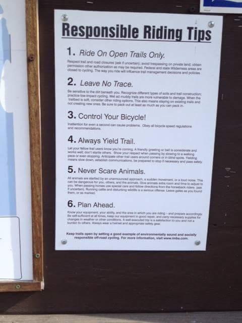 A bulletin board displaying a list of responsible riding tips for cyclists, outlining six key guidelines: 1. Ride on open trails only, 2. Leave no trace, 3. Control your bicycle, 4. Always yield trail, 5. Never scare animals, and 6. Plan ahead. The tips are intended to promote safe and environmentally conscious cycling practices. Hueston Woods State Park mountain bike trail.