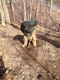 A person using a shovel to dig in a forested area, surrounded by trees and fallen leaves, on a sunny day. Hueston Woods State Park mountain bike trail.