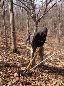 Two individuals are working in a wooded area during a cool season, one in the foreground is digging into the ground with a shovel while wearing a hooded sweatshirt, and the other person in the background is also engaged in a similar activity. The scene includes bare trees and fallen leaves, suggesting an outdoor labor project. Hueston Woods State Park mountain bike trail.