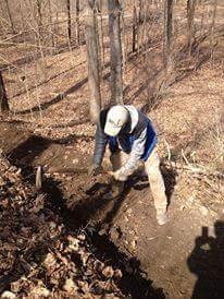 A person wearing a cap and work clothes is digging in a forested area, surrounded by fallen leaves and trees. The scene is set in a natural environment, with sunlight filtering through the branches. Hueston Woods State Park mountain bike trail.