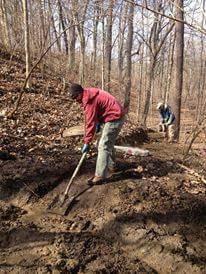 A person digging in a wooded area, wearing a red jacket and green pants, while another individual is seen working in the background. The ground is covered in brown soil and fallen leaves, with trees surrounding the scene. Hueston Woods State Park mountain bike trail.