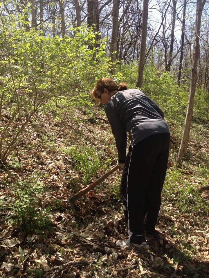 A person wearing a black athletic outfit is bent over in a wooded area, using a digging tool to work in the ground covered with fallen leaves. Sunlight filters through the trees, highlighting the greenery around. Hueston Woods State Park mountain bike trail.