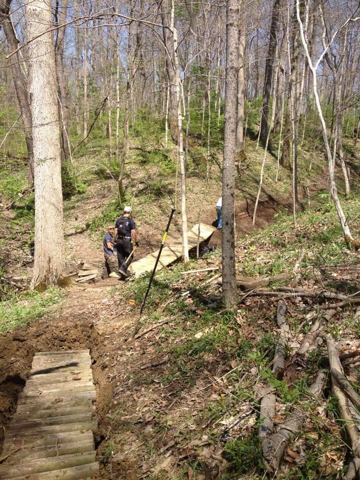 Two individuals working on a hiking trail in a forested area. One is using a tool to level the ground while the other stands nearby, both surrounded by trees and underbrush. Wooden planks are laid out to create a pathway. The scene is set under a clear blue sky, indicating a sunny day. Hueston Woods State Park mountain bike trail.