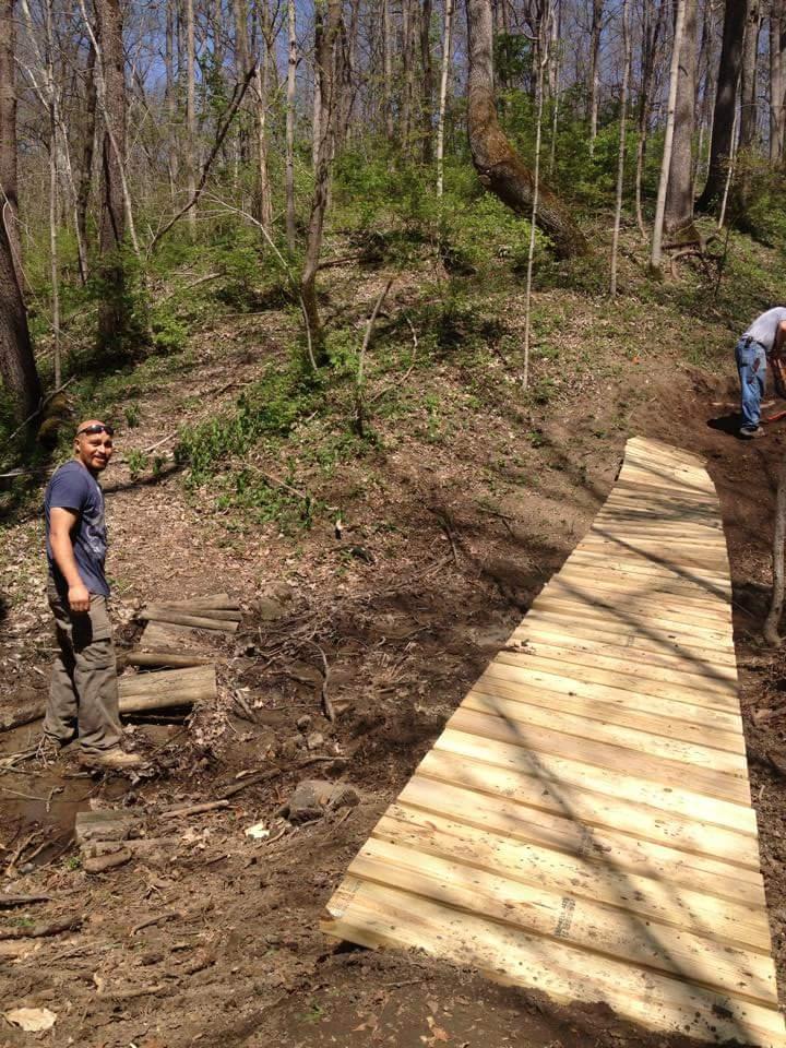 Two people working on constructing a wooden trail in a forested area. One person stands on the left, smiling and wearing a blue shirt, while another person is visible in the background, engaged in the construction. The wooden path is laid out, extending along the dirt ground, surrounded by trees and greenery under a clear blue sky. Hueston Woods State Park mountain bike trail.
