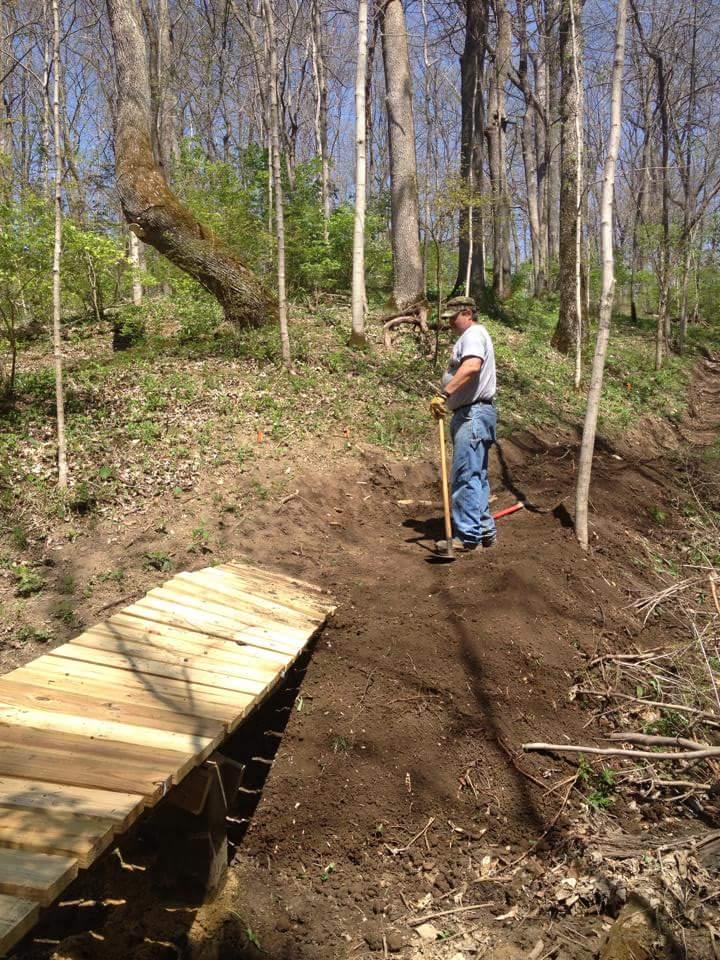 A person standing on a dirt path in a wooded area, wearing a t-shirt and jeans, holding a shovel. Nearby, a wooden bridge is built over the ground, surrounded by trees and greenery under a clear blue sky. Hueston Woods State Park mountain bike trail.