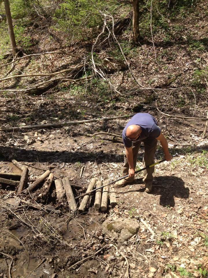 An individual working in a wooded area, using a long stick to adjust logs on the ground. The surrounding environment features green foliage, fallen branches, and a muddy patch. Hueston Woods State Park mountain bike trail.