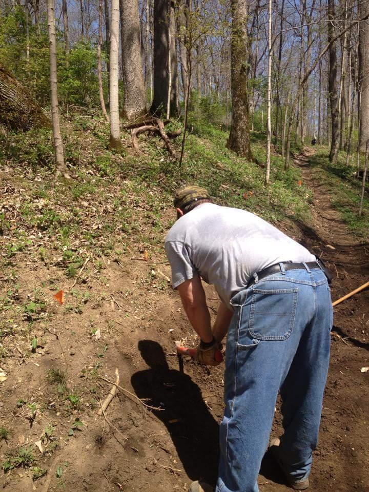 A person dressed in a gray t-shirt and blue jeans is kneeling on the ground in a forested area, focusing on digging into the soil with a tool. The background features tall trees and a dirt path winding through the greenery, indicating a natural outdoor setting. Bright sunlight illuminates the scene, highlighting the vibrant foliage. Hueston Woods State Park mountain bike trail.