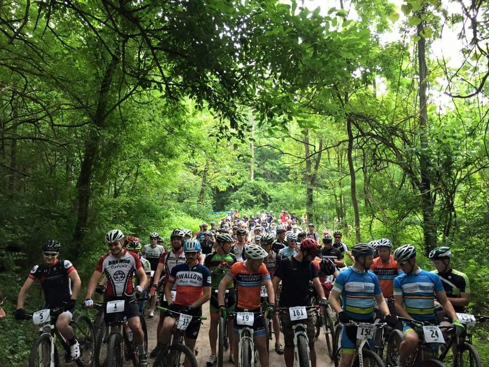 A large group of mountain bikers gathered on a dirt trail surrounded by dense green foliage. The cyclists, wearing various brightly colored jerseys and helmets, are preparing for a race. The scene captures the excitement and camaraderie of the event in a lush, natural setting. Hueston Woods State Park mountain bike trail.