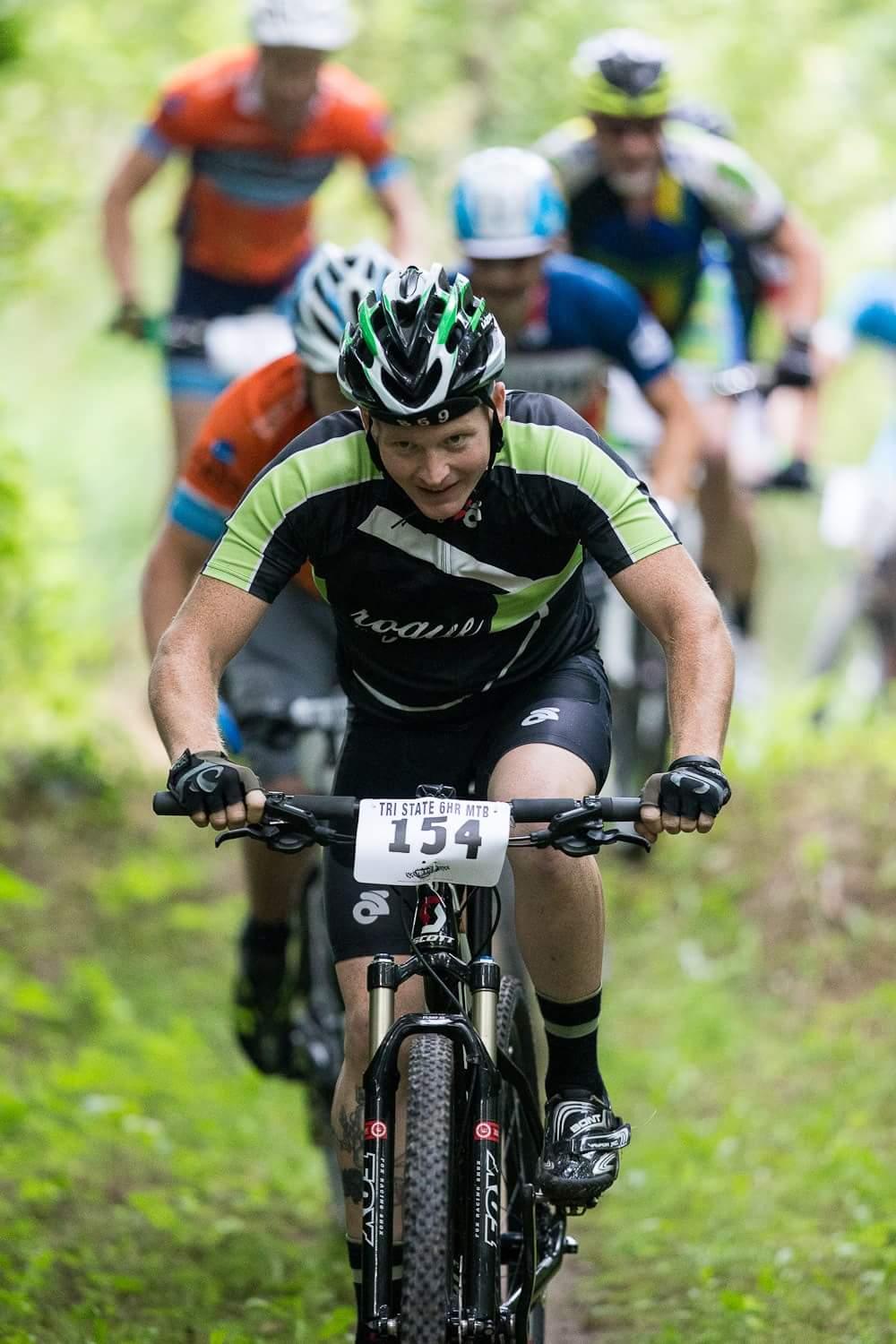 A male mountain biker in a black and green cycling jersey and helmet competes on a dirt path, leading a line of cyclists in the background. The scene is surrounded by lush greenery, indicating a natural, outdoor setting appropriate for a mountain biking event. The cyclist is focused and appears determined as he pedals forward, with a race number displayed on his bike. Hueston Woods State Park mountain bike trail.