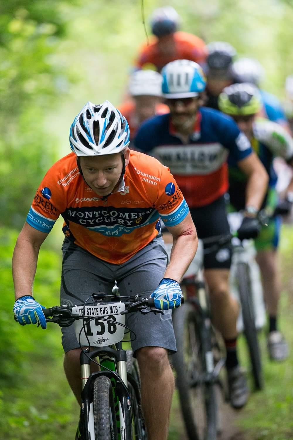 A group of mountain bikers competing in a race, with a focus on one rider in an orange jersey and blue gloves. The cyclist is leaning forward on their bike, navigating a trail surrounded by greenery, while others follow behind. The atmosphere captures the intensity of the event. Hueston Woods State Park mountain bike trail.