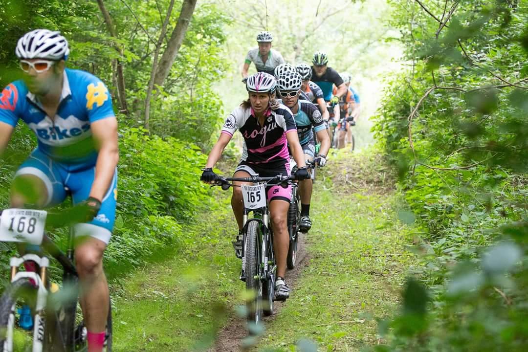 A group of mountain bikers riding along a narrow trail surrounded by lush greenery. The lead cyclist, wearing a black and pink jersey with the number 165, is followed closely by others in colorful cycling attire. The scene conveys an active and competitive atmosphere in a natural setting. Hueston Woods State Park mountain bike trail.