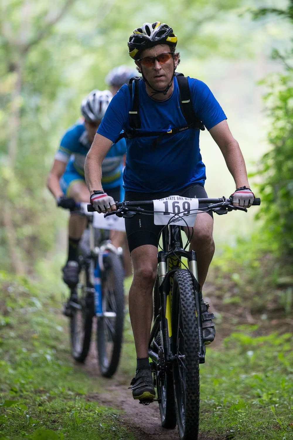 A mountain biker wearing a blue shirt and helmet rides along a dirt trail, focused on the path ahead. In the background, another cyclist in a blue and white outfit follows closely. The scene is surrounded by lush greenery, indicating a natural outdoor setting. The cyclist has a race number visible on their shorts. Hueston Woods State Park mountain bike trail.