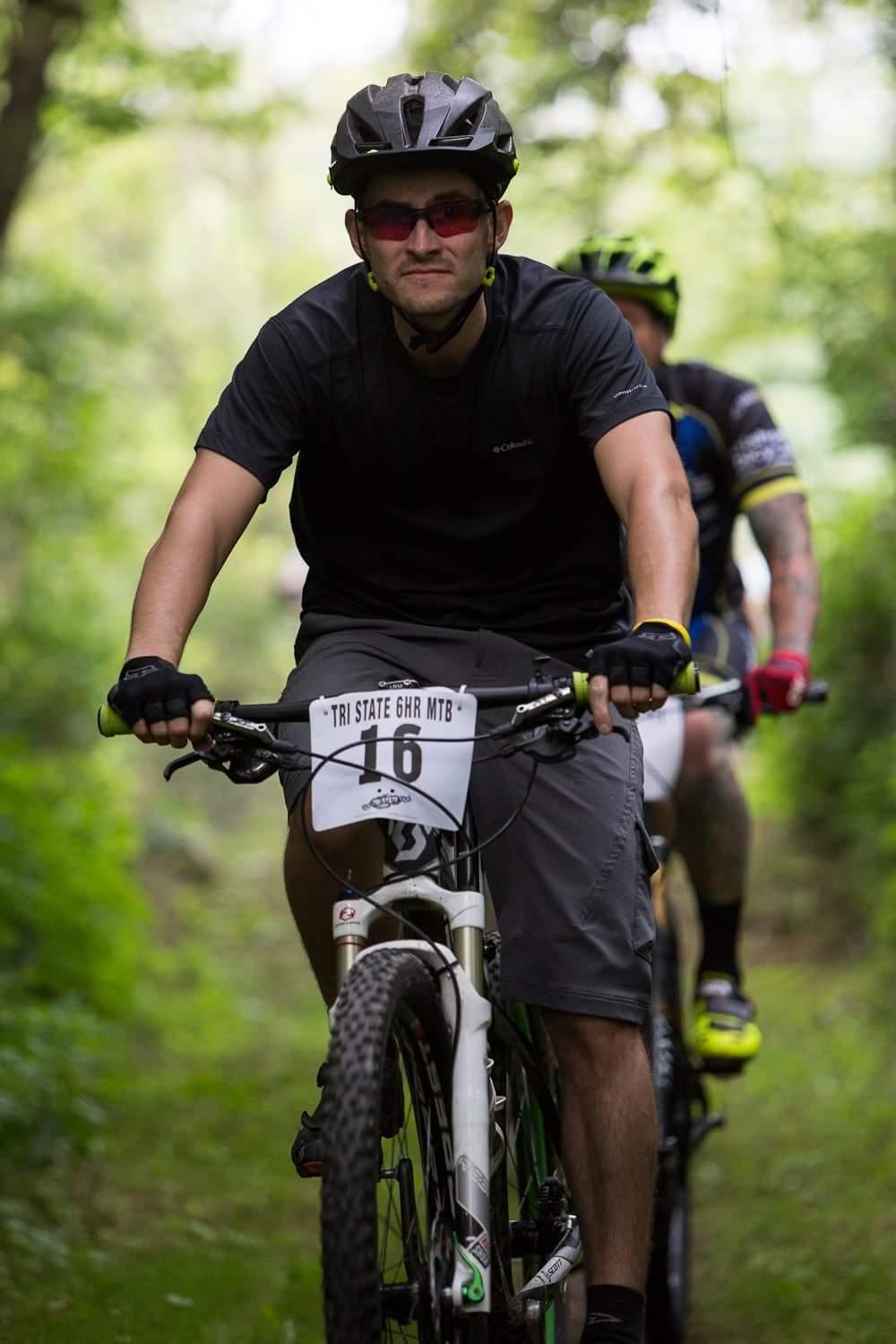A mountain biker wearing a black helmet and sunglasses rides on a forest trail. He is focused, with his hands gripping the handlebars of a white mountain bike, which has a race number labeled "16" attached to the front. In the background, another cyclist is seen, slightly blurred, contributing to the atmosphere of an outdoor biking event. The lush greenery of the surrounding trees adds to the natural setting. Hueston Woods State Park mountain bike trail.