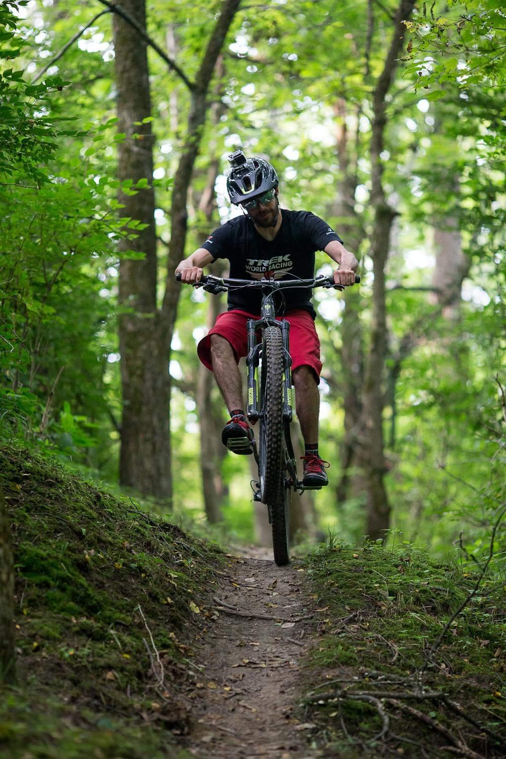 A person riding a mountain bike on a narrow trail in a lush green forest, performing a wheelie. The rider is wearing a helmet and a black t-shirt with a logo, paired with red shorts. Surrounding trees and foliage create a vibrant, natural backdrop. Hueston Woods State Park mountain bike trail.