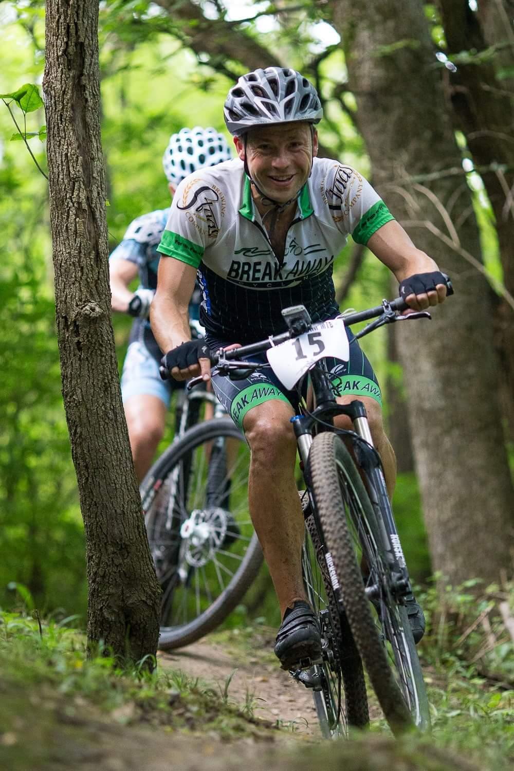 A mountain biker navigating a wooded trail, smiling as he rides. He is wearing a cycling jersey with the text "BREAK AWAY" and has a helmet on. Another cyclist is visible in the background, also riding a mountain bike. The scene is lush with green foliage and trees. Hueston Woods State Park mountain bike trail.