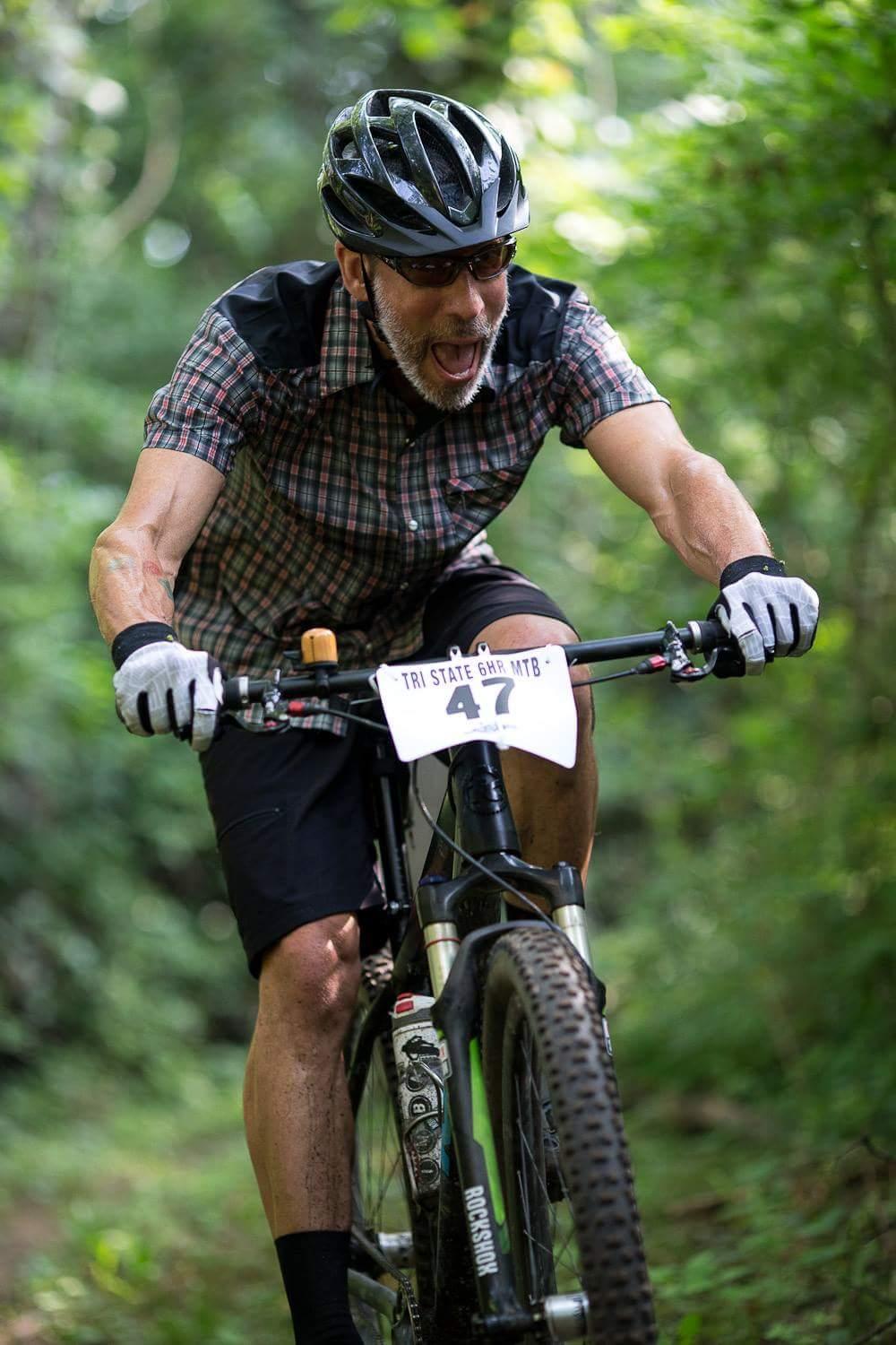 A cyclist wearing a helmet and sunglasses rides vigorously on a mountain bike along a lush, wooded trail. He is dressed in a checkered shirt and shorts, and his expression reflects excitement and focus. The bike number tag on the front indicates participation in a mountain biking event. The background features blurred greenery, creating a vibrant outdoor atmosphere. Hueston Woods State Park mountain bike trail.