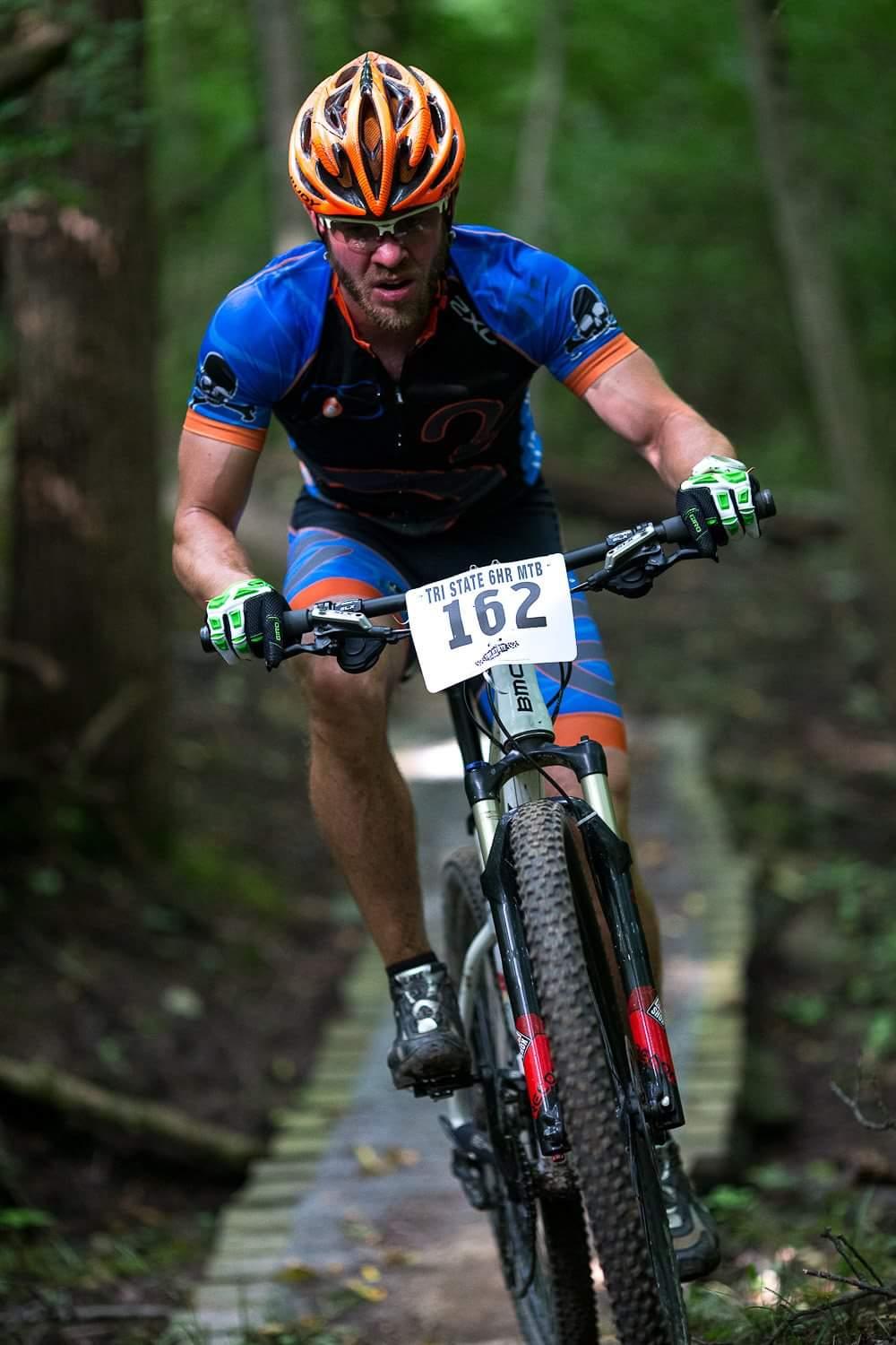 A mountain biker navigating a narrow wooden trail in a forest. The cyclist is wearing a blue and orange jersey and helmet, with a race number displayed on the bike. The bike has thick tires suitable for rough terrain, and the background features lush green trees. The rider looks focused and determined as he pedals forward. Hueston Woods State Park mountain bike trail.