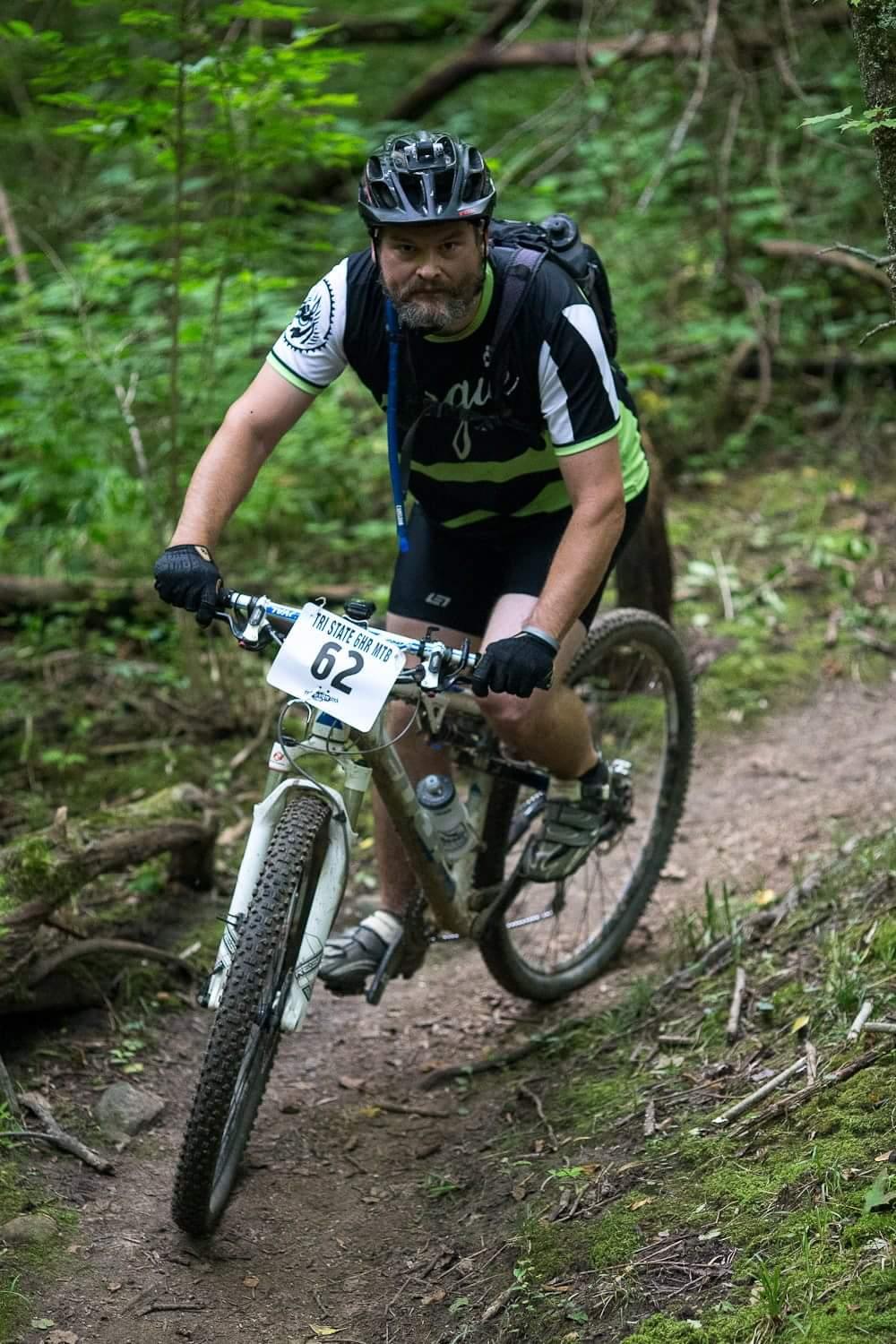 A man riding a mountain bike on a wooded trail, wearing a helmet and sporting a cycling jersey with green and black stripes. He is focused and navigating a bend in the path, with a race number pinned to his bike. The surrounding area is lush with greenery, indicating a scenic outdoor environment. Hueston Woods State Park mountain bike trail.