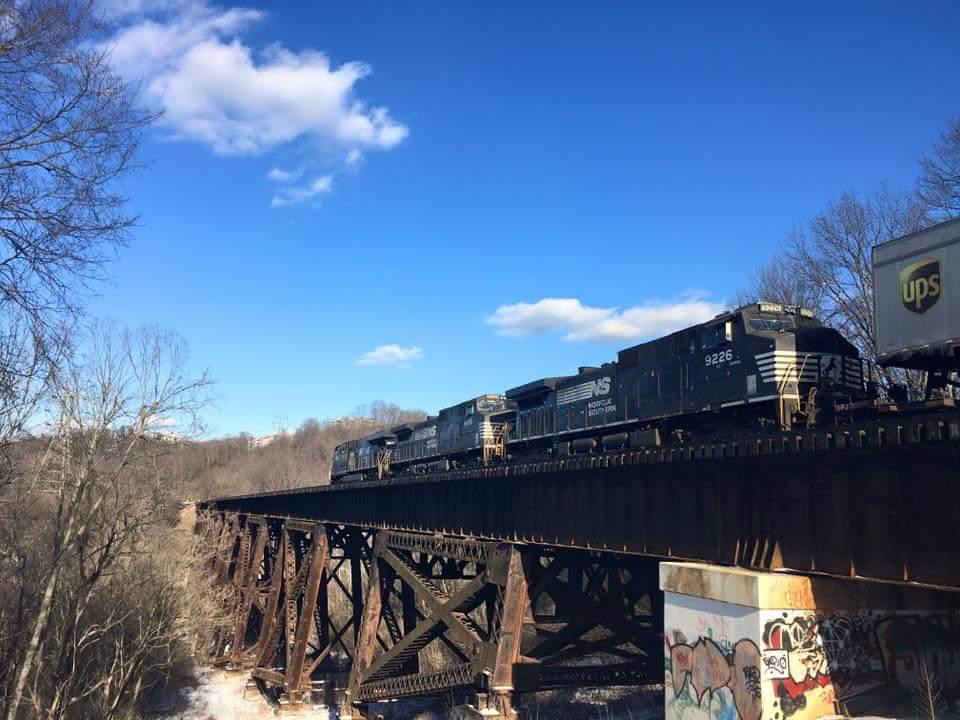 A freight train consisting of multiple locomotives travels across a metal trestle bridge. The scene is set against a blue sky with a few clouds, and bare trees line the river below the bridge. Graffiti is visible on the bridge structure, and a UPS truck can be seen in the foreground. Devou Park mountain bike trail.