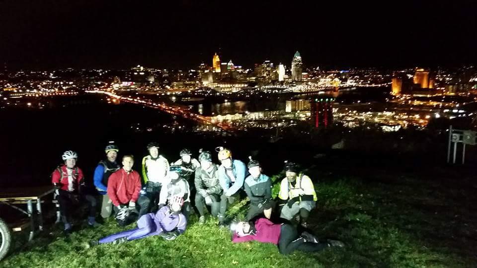 A group of cyclists posing together on a grassy hill at night, with a city skyline illuminated in the background. Some participants are sitting, while others are lying down, all smiling and wearing cycling gear and helmets. The city lights reflect off the water, creating a vibrant nighttime atmosphere. Devou Park mountain bike trail.