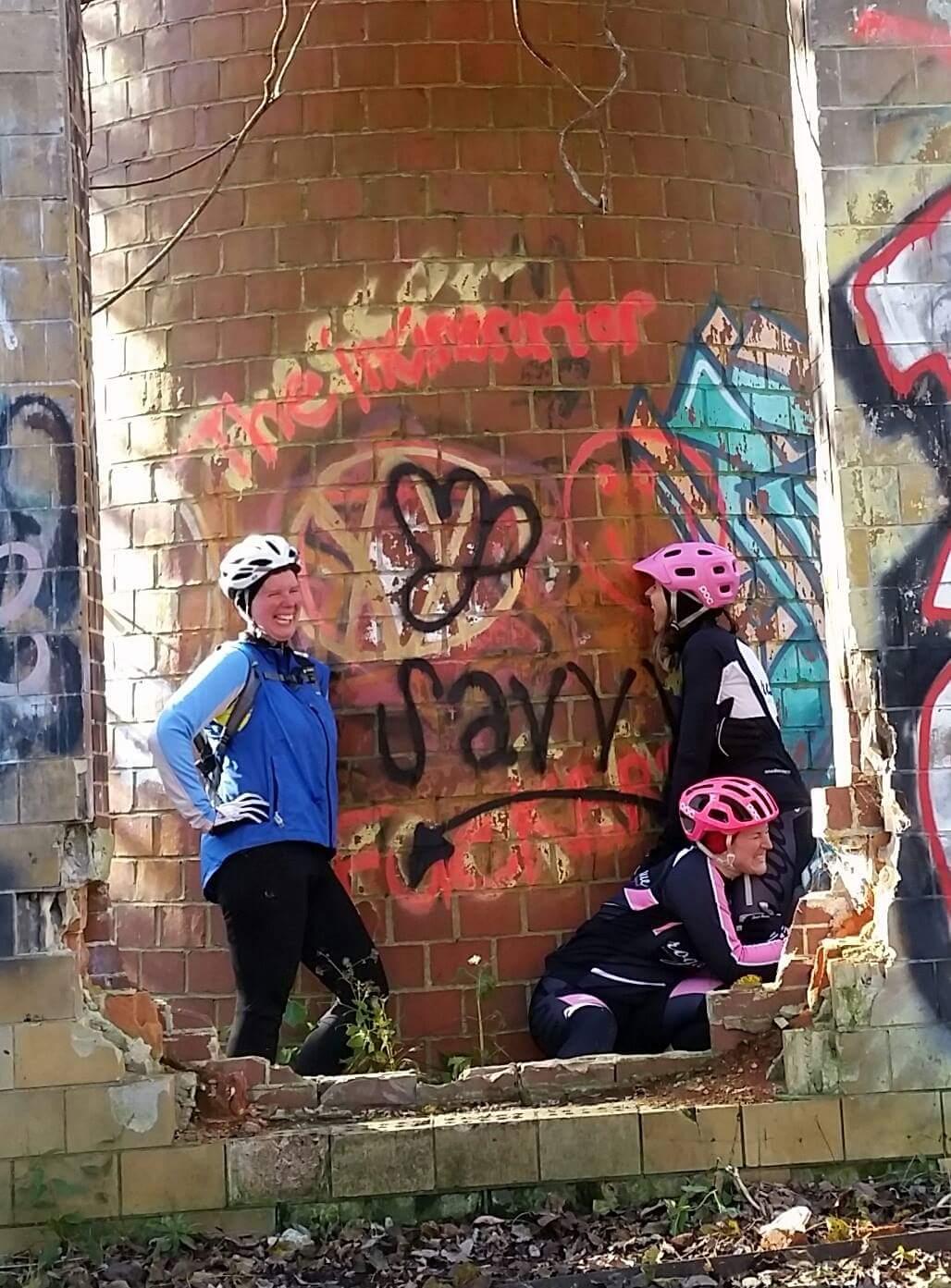 Three women in cycling attire pose playfully beside a graffiti-covered brick pillar. One woman stands confidently with her hands on her hips, while another leans against the pillar, and the third crouches playfully beside them. They are all wearing helmets and smiling, set against a backdrop of urban art and a partially damaged wall. Green foliage peeks through the cracks in the brick structure, indicating an outdoor setting. Devou Park mountain bike trail.