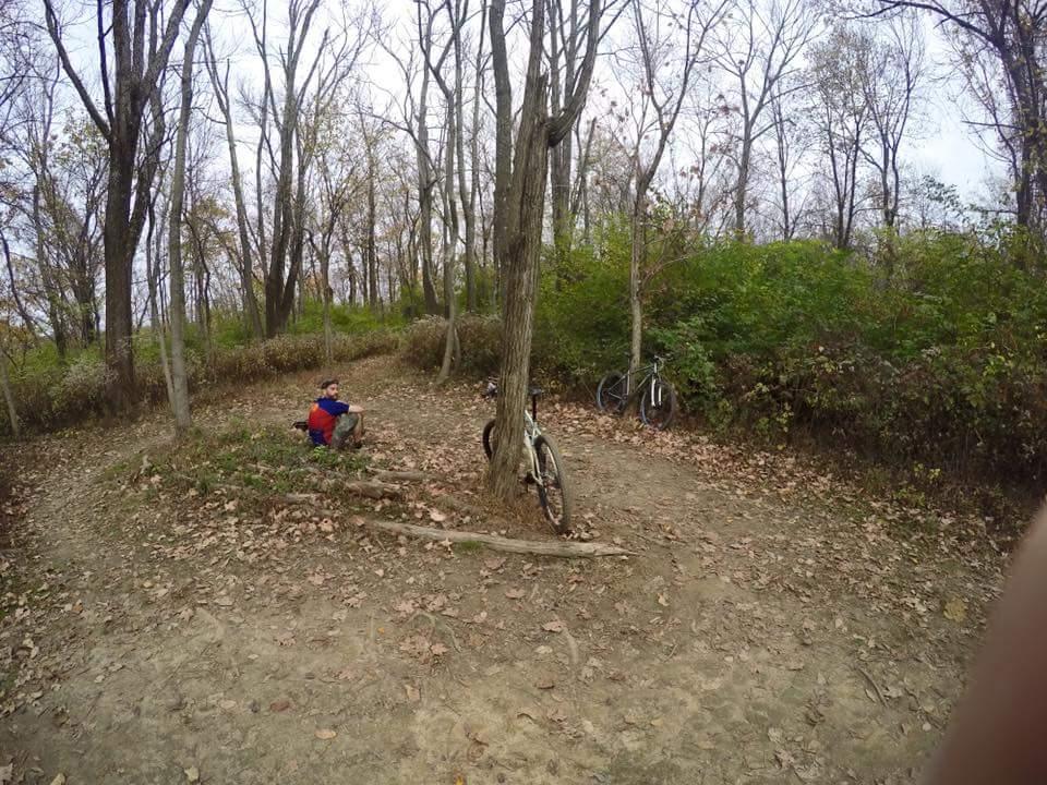 A young person sitting on the ground next to a tree in a wooded area, surrounded by autumn leaves. Two bicycles are parked against the tree, and the scene features bare trees and greenery in the background under a cloudy sky. Devou Park mountain bike trail.