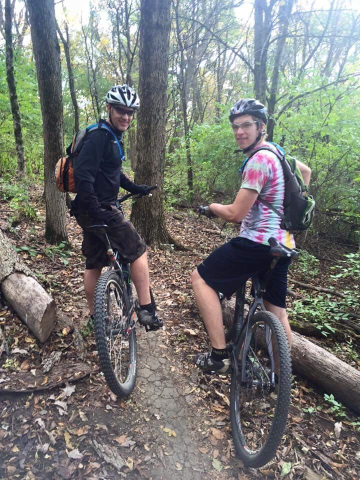 Two young men on mountain bikes pause on a dirt trail surrounded by trees. One is wearing a black helmet and a black shirt with shorts, while the other has a colorful tie-dye shirt, a black helmet, and a backpack. They are both smiling and facing slightly toward the camera, with greenery and logs visible in the background. Devou Park mountain bike trail.