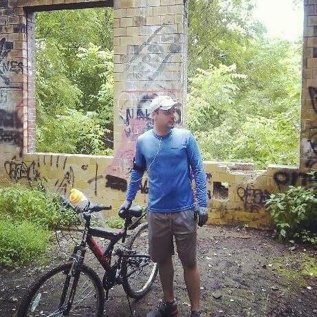 A person standing near a graffiti-covered wall inside a ruined structure, holding a bicycle. The scene is surrounded by lush green foliage, indicating a forested area. The individual is wearing a blue long-sleeve shirt, shorts, and a cap. Devou Park mountain bike trail.