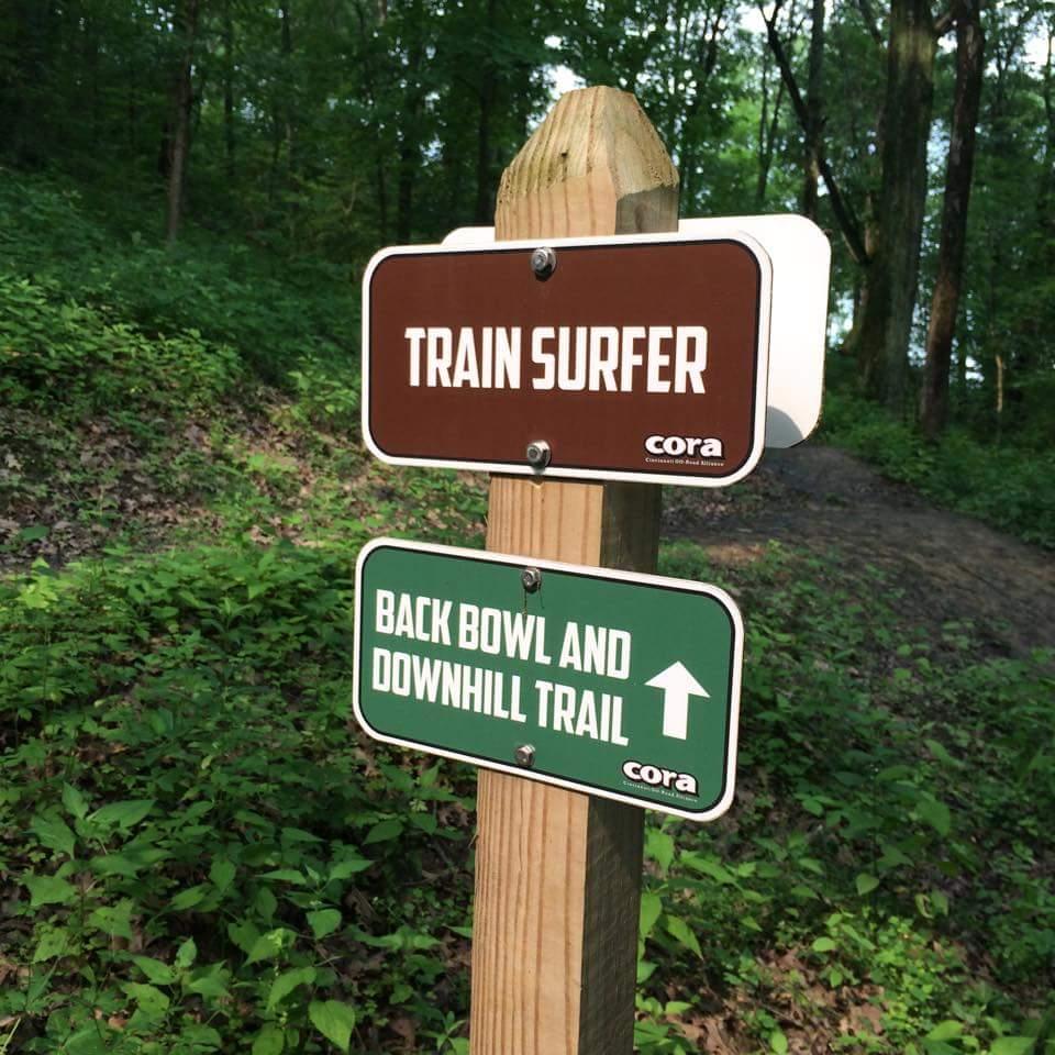 Signpost in a wooded area displaying two trail markers. The top sign reads "TRAIN SURFER" in white letters on a brown background, while the bottom sign indicates "BACK BOWL AND DOWNHILL TRAIL" with an arrow, in white letters on a green background. The surrounding landscape features lush greenery. Devou Park mountain bike trail.