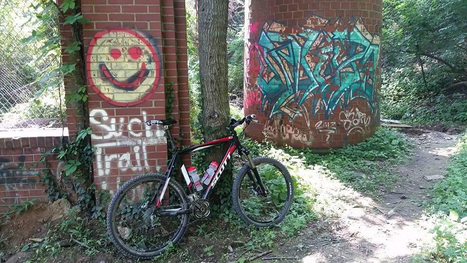 A mountain bike is leaning against a brick wall covered in graffiti, featuring a smiley face and the phrase "Such Trail." In the background, another brick structure with blue and green graffiti can be seen, surrounded by greenery and a dirt path leading through the area. Devou Park mountain bike trail.