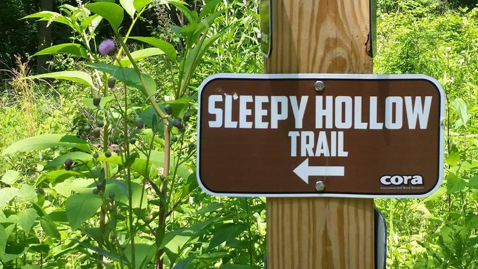 Sign marking the "Sleepy Hollow Trail" with an arrow pointing to the left, surrounded by green vegetation. Devou Park mountain bike trail.