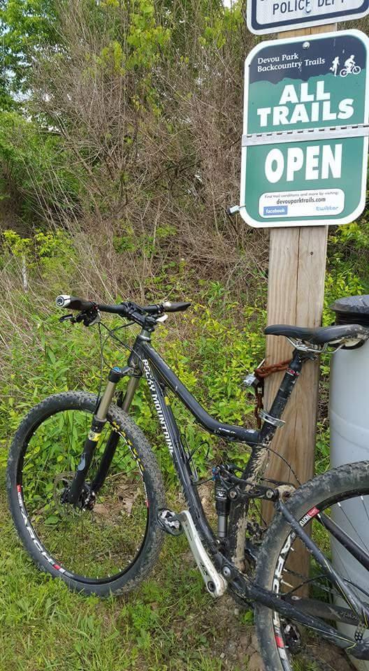 A mountain bike resting against a wooden sign that reads "All Trails Open" at Devout Park Backcountry Trails. The bike is covered in dirt, indicating recent use, and the surrounding area features lush greenery. A trash can is visible nearby, contributing to the outdoor trail environment. Devou Park mountain bike trail.