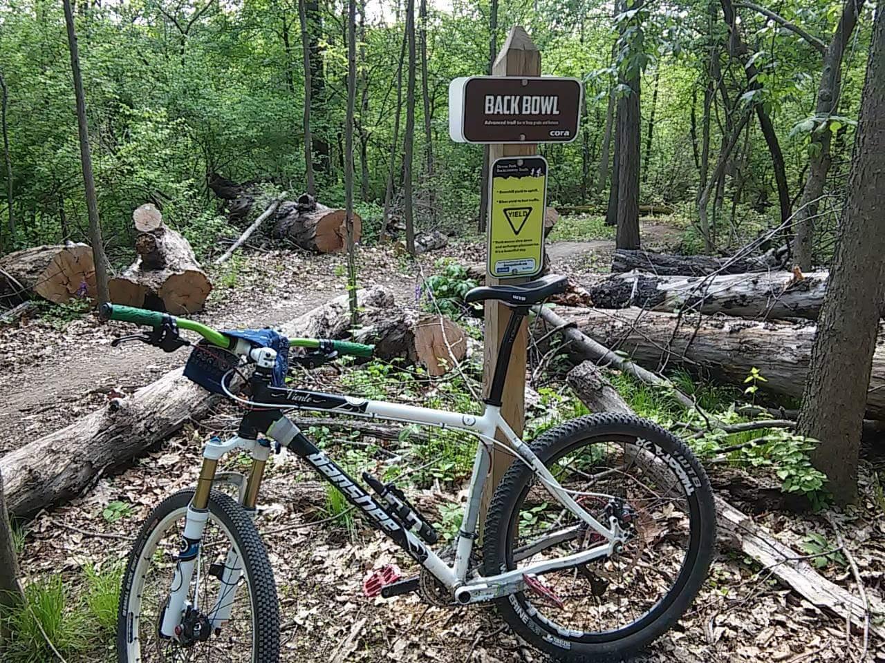 A mountain bike parked beside a trail sign labeled "Back Bowl" in a wooded area. The path is surrounded by fallen logs and dense greenery, indicating an advanced biking trail. Devou Park mountain bike trail.