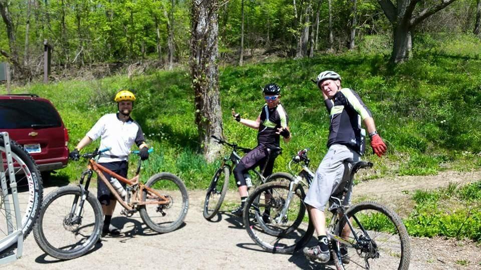 Three mountain bikers pose with their bikes in a sunny outdoor setting, surrounded by greenery. They are wearing helmets and biking gear, smiling and making hand gestures. A red vehicle is parked in the background. Devou Park mountain bike trail.
