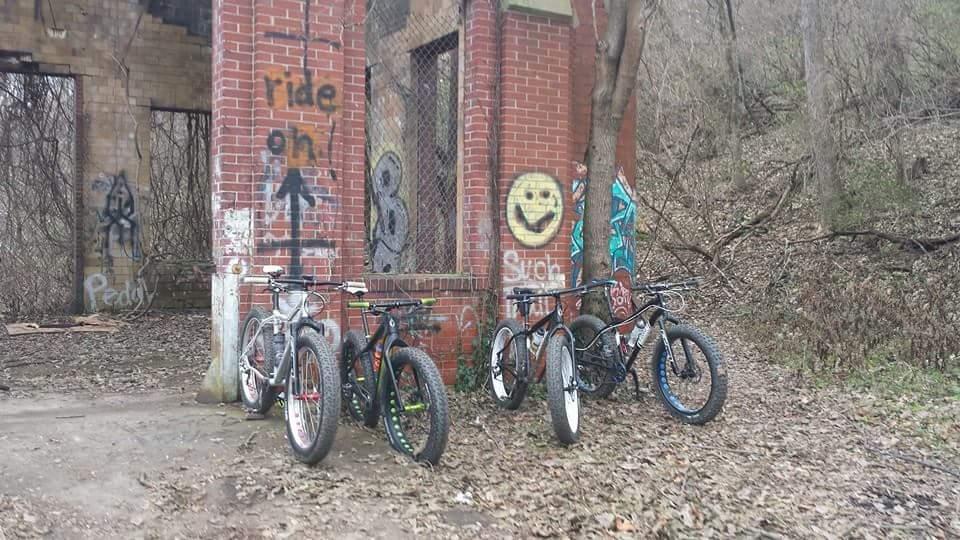 A group of bicycles parked next to a brick wall covered in graffiti, with a smiling face and the words "ride on" visible. The ground is littered with leaves, and the surrounding area is wooded. Devou Park mountain bike trail.