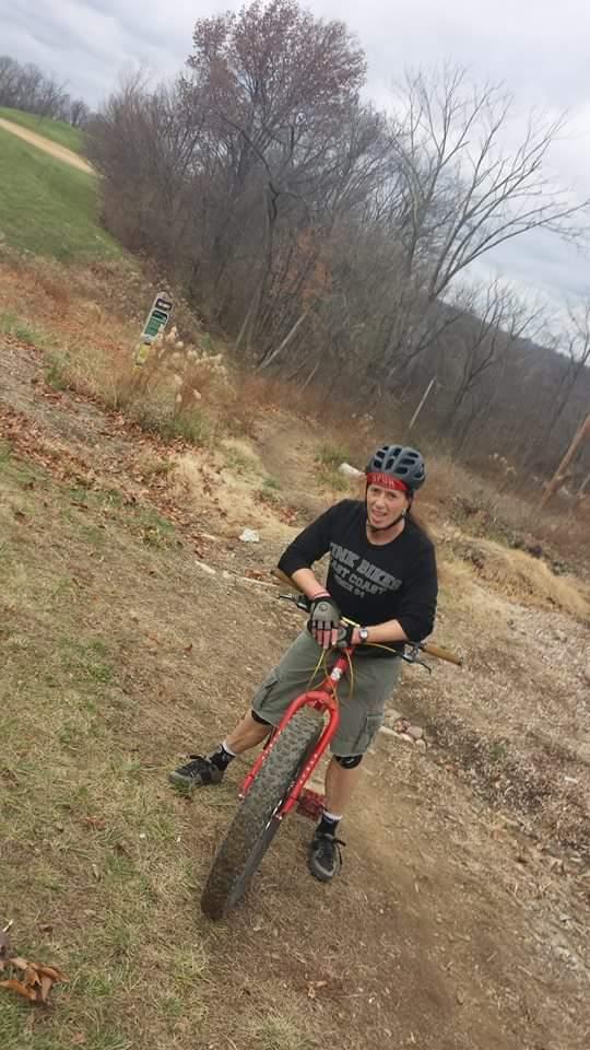 A person wearing a black helmet and gloves stands next to a red mountain bike on a dirt trail. They are dressed in a black long-sleeve shirt and gray shorts, smiling at the camera. The background features a cloudy sky, bare trees, and a grassy area, suggesting a cool, outdoor environment for biking. Devou Park mountain bike trail.