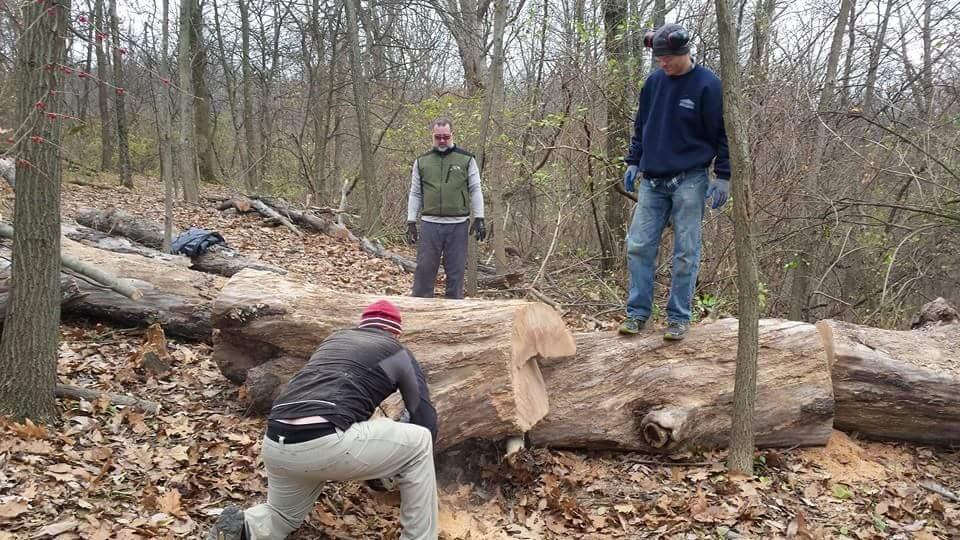 Three men are working in a wooded area. One man, dressed in a dark jacket and a red beanie, is crouched next to a large log, using a tool to carve or chop it. Another man stands on top of the log, wearing a dark sweatshirt and gloves, while the third man observes nearby, dressed in a green vest. The ground is covered in fallen leaves and there are other logs in the background, typical of a forest environment. Devou Park mountain bike trail.